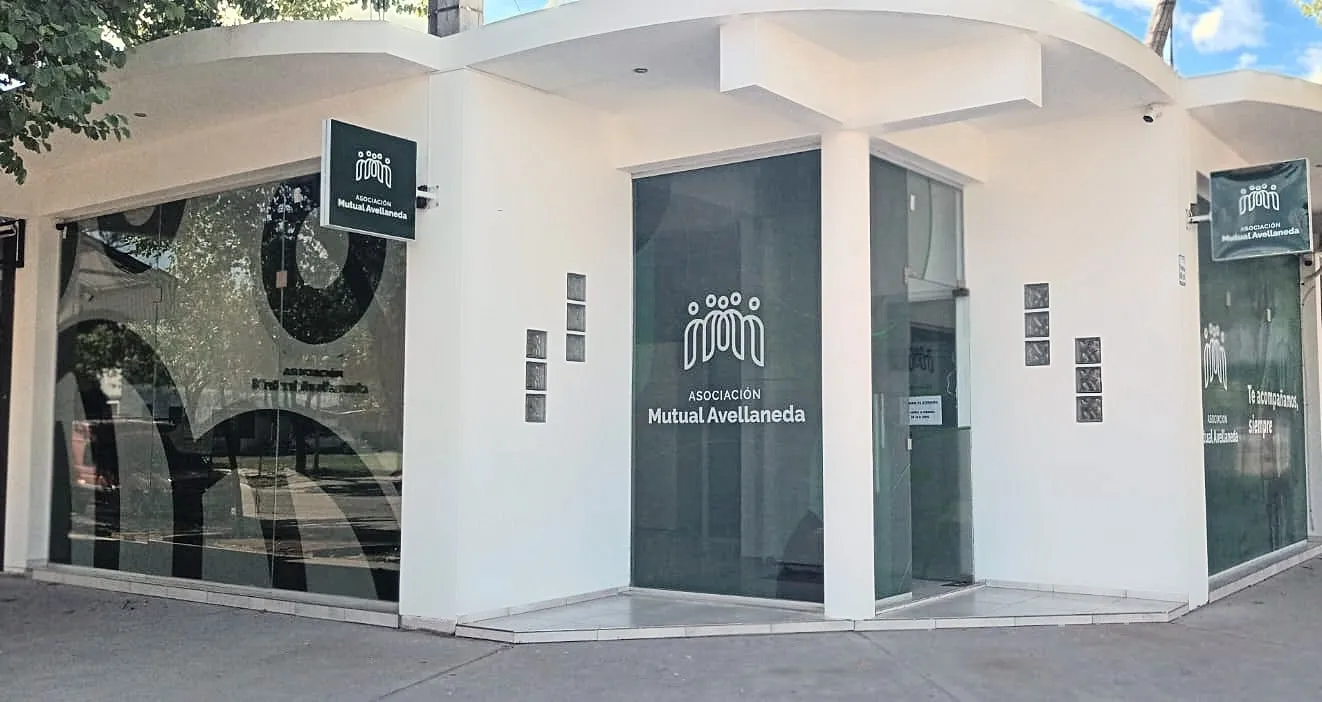 Frente de la sede de la Asociación Mutual Avellaneda: local blanco en esquina, con el logo en los vidrios y en dos carteles, y el lema «Te acompañamos, siempre»